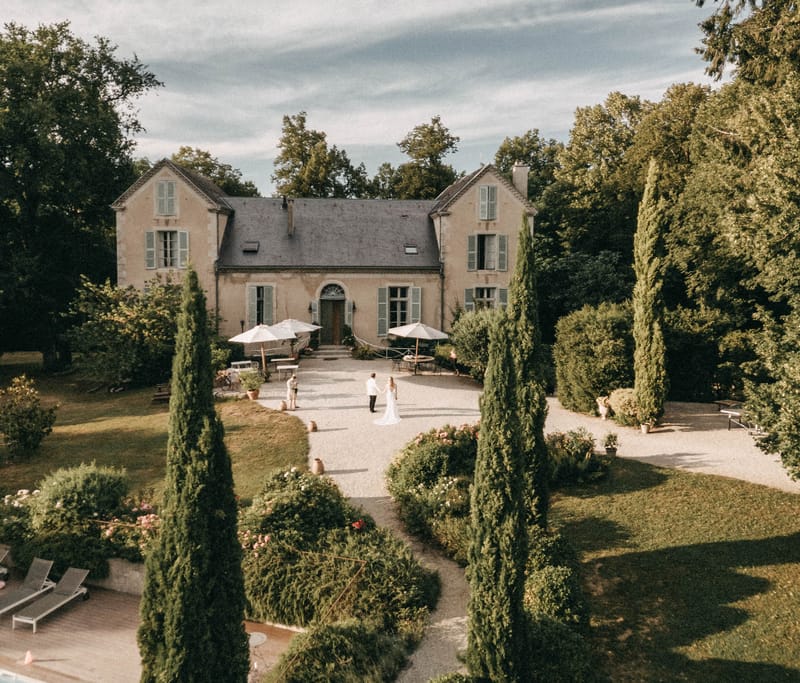 Aerial drone view of couple on gravel courtyard of a French manor house with cypress trees, hedgerows, and pool area