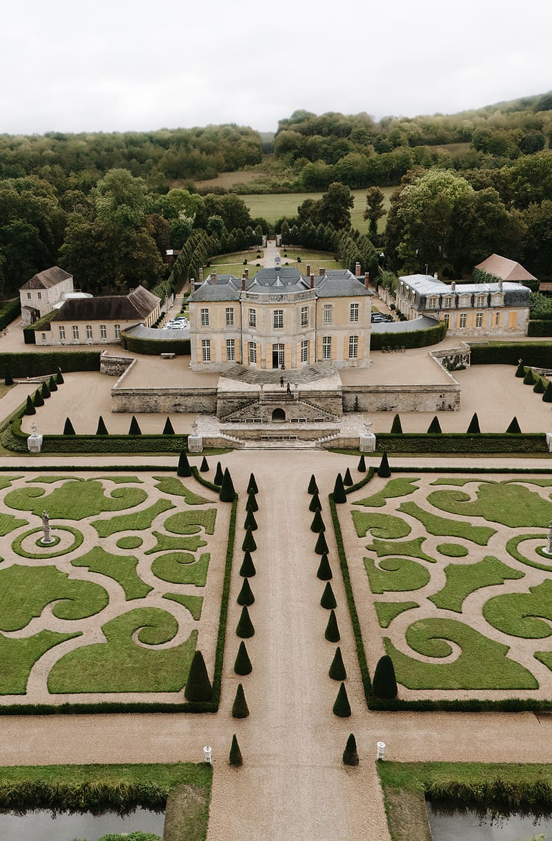 An aerial drone shot looking down the central axis of a French château and its formal French-style parterre gardens. The château is a classic 18th-century-style stone building with a mansard slate roof, symmetrical facades, and flanking outbuildings on either side. The foreground is dominated by intricately clipped boxwood parterres in scrollwork patterns, flanked by rows of cone-shaped topiary trees lining a gravel central allée. Two small figures, likely the couple, are visible on the stone staircase at the entrance of the château, providing a sense of scale. The wider grounds show hedgerow borders, a long driveway leading away from the rear of the château, and dense woodland beyond. Potential venue feature image.
