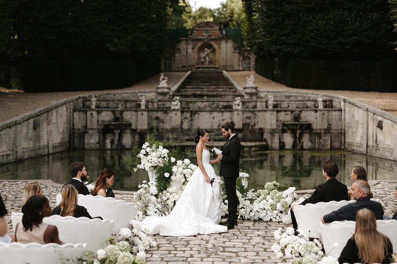 An outdoor wedding ceremony taking place on a cobblestone terrace in front of a grand ornamental stone fountain basin with classical statuary and a symmetrical staircase backdrop. The bride wears a strapless white ballgown with a full skirt and cathedral-length train, while the groom is dressed in a black tuxedo with bow tie and his hair in a bun; he is reading from a paper, likely his vows. The ceremony altar is flanked by large white urns overflowing with white peonies, ranunculus, and ferns, with additional ground-level arrangements of white blooms — including what appears to be baby's breath and hydrangea — lining the aisle. Guests, dressed predominantly in black formal attire, are seated in white ornate carved wooden chairs arranged on either side of the aisle, with approximately 20–30 visible guests. The overall styling is classic and monochromatic, with a strict white and green floral palette against the grey stone architecture. This is a wide editorial shot that frames the couple centrally with the full ceremony setting and architectural backdrop visible. Potential venue feature image.