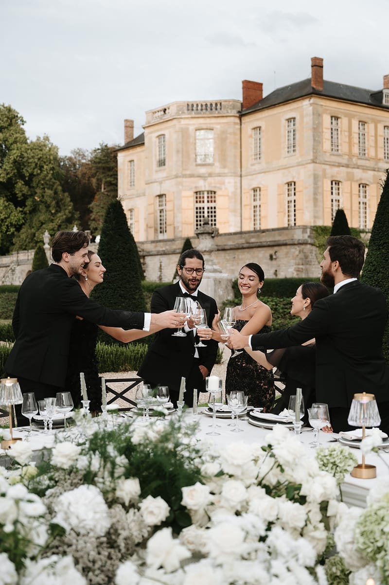A group of six guests raises wine glasses in a toast during an outdoor reception dinner at a French château. The guests are dressed in an all-black dress code — the men in black tuxedos and the women in black evening gowns, one featuring a lace overlay. The rectangular dining table is set with white linen, dark charger plates, crystal glassware, white pillar candles, small brass accent lamps, and a lush floral runner composed of white garden roses, white hydrangeas, and green foliage. The formal French garden setting features clipped topiary cones and hedges, with a large honey-toned stone château visible in the background. The overall decor palette is black, white, and gold with a formal, modern classic aesthetic. Medium wide shot taken from a slightly low angle at table level, with the château centered in the background. Potential venue feature image.