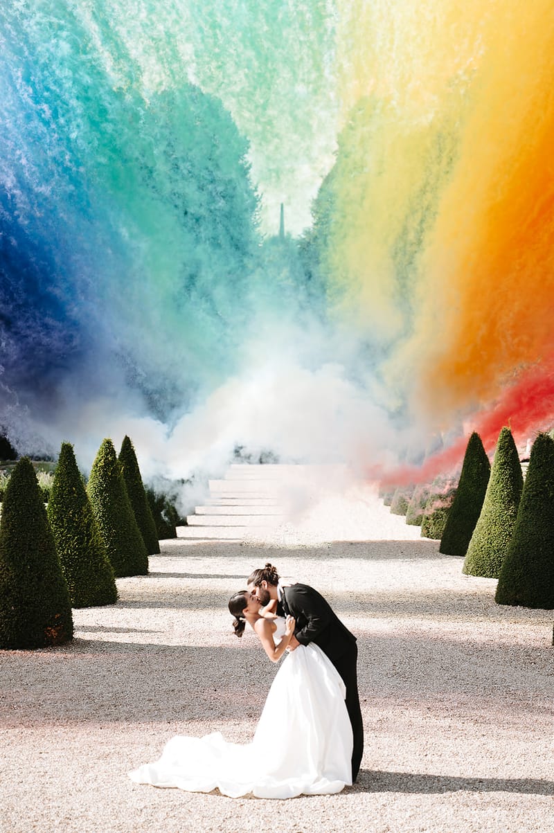 A couple shares a dip kiss during an outdoor portrait session on a gravel allée lined with cone-shaped topiary trees, with white stone steps leading upward in the background. The groom wears a black suit and dips the bride, who is dressed in a white ballgown with a full skirt and trailing train, her hair styled in an updo. A large-scale rainbow smoke bomb display fills the upper portion of the frame, with vivid plumes of blue, teal, green, yellow, orange, and red smoke billowing above and around the couple against the formal garden setting. The wide-shot composition centers the couple at the base of the allée, using the row of topiaries and receding path as leading lines into the colorful smoke backdrop.