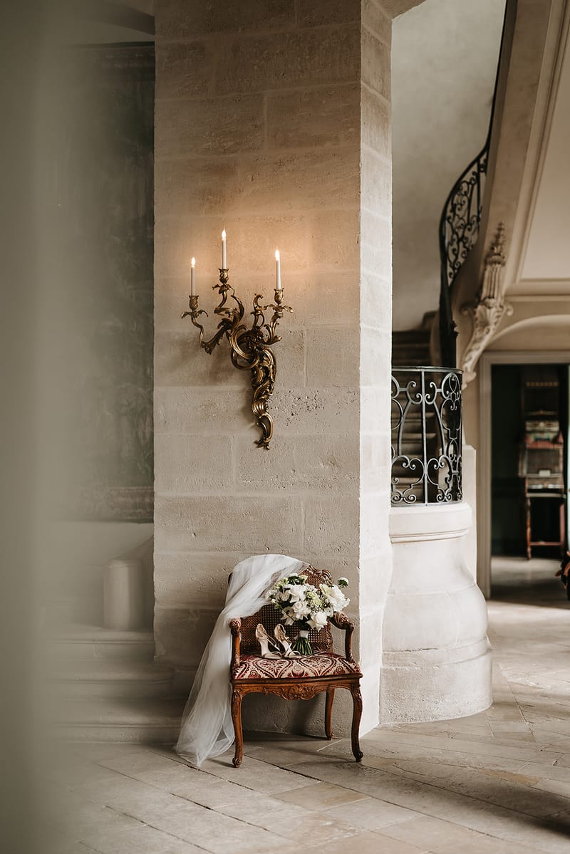 A bridal detail flat lay styled on an antique French armchair with a red and gold patterned upholstered seat, positioned against a cut-stone wall inside a château. The chair holds a bouquet of white roses and green foliage, a pair of nude strappy heeled sandals, and a long white tulle veil that drapes down to the stone tile floor. Above the chair, a gilded bronze wall sconce with an ornate rococo design holds two lit white taper candles. To the right, an iron spiral staircase with decorative scrollwork is partially visible, along with a glimpse into an adjoining room with dark green walls. The composition is a wide environmental shot that places the bridal accessories within the architectural setting of the venue interior. Potential venue feature image.