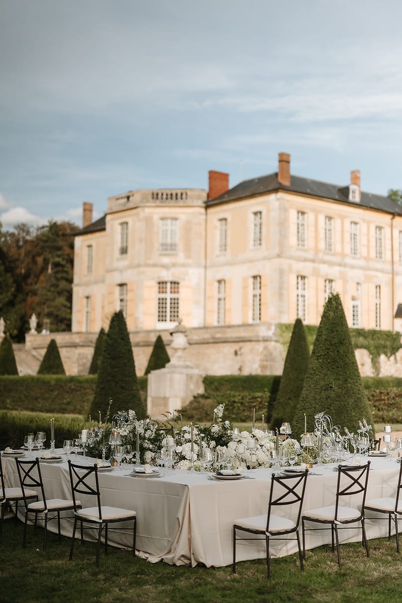 An outdoor wedding reception table is set in the formal garden of a French château, with the pale stone manor house and its conical topiary trees visible in the soft-focus background. The large oval dining table is dressed in an ivory linen tablecloth and surrounded by dark metal cross-back chairs with cream cushioned seats. The centerpiece is a lush runner of white florals — including what appear to be white roses, ranunculus, and orchids — interspersed with greenery and delicate trailing botanicals, running the full length of the table. Thin gold taper candles in individual holders are placed throughout the arrangement alongside crystal glassware, white charger plates, and scattered votive candles, creating a classic, white-and-green décor palette. Wide shot composition with the table in the foreground and the château grounds receding into the background. Potential venue feature image.