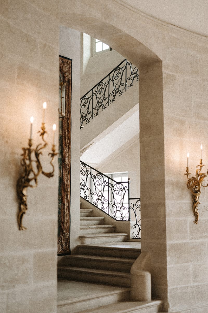 An interior architectural detail shot of a grand staircase viewed through a stone archway, with no people present. The staircase features ornate black wrought-iron scrollwork balustrades and pale stone steps that curve upward toward a tall window. Two gilded brass candle-style wall sconces flank the archway on either side, each fitted with lit taper candles that cast a warm glow against the cream-colored cut-stone walls. A decorative gilt-framed panel or tapestry fragment is partially visible on the left side of the arch. The composition is a medium interior shot framed by the arch, emphasizing the classic French château architectural style. Potential venue feature image.
