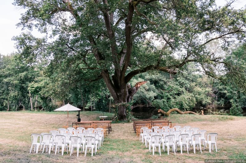 An outdoor ceremony setup photographed before guests arrive, arranged on an open lawn in front of a large, mature oak tree with a small body of water visible behind it. Seating is arranged in two sections divided by a central aisle, consisting of white plastic garden chairs in the outer rows and darker wooden benches closer to the front. A simple wooden altar or table is positioned at the head of the aisle, with a cream-colored parasol on a stand placed to one side, likely providing shade for the officiant. The overall styling is informal and rustic, with no visible floral arch or decorative ceremony backdrop. Wide shot capturing the full ceremony layout and surrounding wooded landscape.