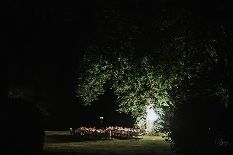 A wide-angle nighttime shot of an outdoor wedding reception dinner set up on a lawn, with multiple long rectangular tables arranged in a U or parallel formation, dressed in white linens and set with glassware, candles, and floral centerpieces in warm amber tones. The tables are surrounded by cross-back wooden chairs and lit by warm candlelight and what appears to be an overhead chandelier or hanging light installation suspended from a large mature tree, which is dramatically uplighted in green. The surrounding grounds are dark, with the illuminated table setting and tree forming a focal point against the black night sky. No guests are visible, suggesting the shot was taken before or between service. The composition is a distant wide shot taken from ground level across the lawn, framing the scene through the dark silhouette of hedgerows.