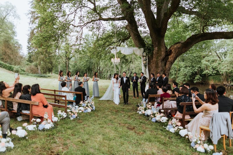 The bride and groom walk back down the aisle together following the conclusion of an outdoor ceremony, held on a grass lawn beneath a large mature tree. The ceremony arch is a white draped teepee-style structure with a crystal chandelier suspended from it, decorated with florals, positioned under the tree canopy. The aisle is lined on both sides with low floral arrangements featuring white ranunculus, blue hydrangeas, blush peonies, and delphinium placed directly on the ground. Guests, approximately 40–50 in total, are seated on wooden benches on either side of the aisle and are clapping or taking photos. The bride wears a fitted white lace gown with a long cathedral veil, and the groom wears a black tuxedo with a bow tie. Five bridesmaids stand to the left in misty sage-blue floor-length dresses holding small bouquets, while four groomsmen in black suits stand to the right. The overall styling is romantic-garden with a soft blue, blush, and white floral palette. Wide shot taken from behind the seated guests looking toward the altar.