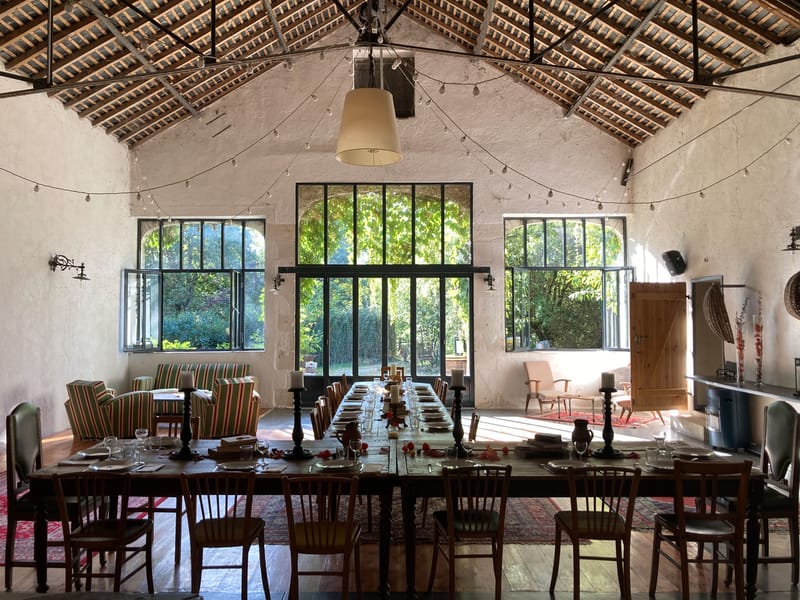 An indoor reception space set up for a wedding dinner, shot as a wide interior view from one end of the room looking toward large black-framed steel and glass windows that open onto a lush green garden. The space has a high vaulted ceiling with exposed wooden beam rafters, white rendered walls, and warm wood plank flooring. Two long rustic wooden farm tables run the length of the room, set with plates, wine glasses, and tall dark-wood candlestick holders with pillar candles; scattered red and orange flower petals and small decorative elements dot the table runners. Mismatched vintage wooden chairs line both tables, contributing to an eclectic, rustic-boho aesthetic. String lights with globe bulbs are draped across the ceiling, and a single large pendant lamp hangs centrally. To the side, a seating area features green and red striped armchairs arranged on a red patterned rug, alongside a cream mid-century armchair, reinforcing the relaxed, lived-in styling. Potential venue feature image.