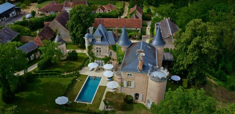 Aerial drone shot of a French château venue featuring multiple conical slate-roofed towers, stone façade with climbing vines, and tall arched windows with white shutters. The property includes a rectangular outdoor swimming pool surrounded by a paved terrace with multiple white market umbrellas and sun loungers, where a small number of people are visible relaxing. The estate complex extends to include several outbuildings with terracotta-tiled roofs arranged around courtyard-style spaces, all set within dense mature tree coverage. No wedding-specific decor or couple is visible in this image. Potential venue feature image.