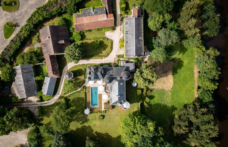 Aerial drone shot of a French country estate complex, showing multiple historic stone and slate-roofed buildings arranged around a central courtyard with a winding gravel driveway. The main manor house features distinctive circular tower elements and is flanked by several outbuildings, barns, and annexes with terracotta and dark slate roofing. A rectangular swimming pool with turquoise water is visible adjacent to the main house, surrounded by white market umbrellas positioned on a paved terrace. The estate grounds include manicured lawns, topiary hedging, and mature trees on all sides. No people or wedding-specific decor are visible in this shot. Potential venue feature image.