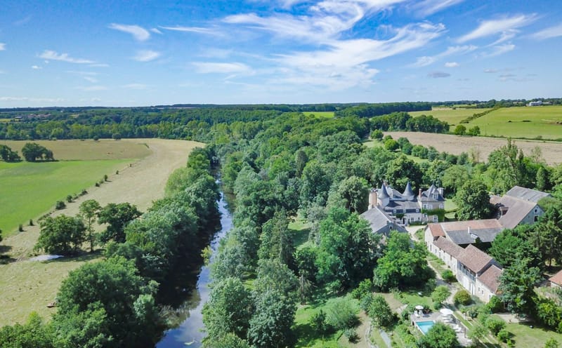 An aerial drone shot showing a French château estate and its surrounding grounds from a wide, elevated perspective. The property features a classic château with pointed turrets and grey slate roofs, accompanied by a cluster of stone outbuildings with terracotta and grey roofing, a rectangular swimming pool with white sun loungers, and a gravel courtyard. The estate sits alongside a calm river bordered by dense tree cover, with open agricultural fields visible on the opposite bank. No people or wedding-specific elements are visible in this image. Potential venue feature image.