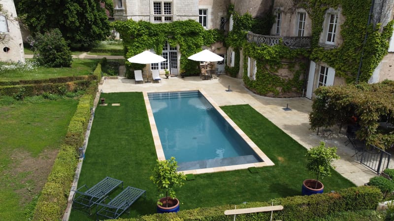 Aerial wide shot of the exterior grounds of a French château venue, showing a rectangular swimming pool with a pale stone surround set within a manicured lawn bordered by trimmed hedges. The château facade is covered in climbing ivy, with tall French windows, a wrought-iron balcony, and white shutters visible. Poolside furniture includes two white market umbrellas, white sun loungers, and dark metal bistro chairs and tables arranged on the stone terrace. Two potted standard trees in cobalt blue ceramic pots are placed near the pool, and additional metal garden benches are positioned at the lower edge of the frame. No people are present. Potential venue feature image.