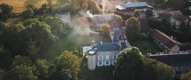 An aerial drone shot taken at golden hour showing a French château estate from above. The main building features a round corner tower with a conical slate roof, blue-shuttered windows, and stone façade construction typical of French country architecture. A small outdoor pool is visible adjacent to the main château. The wider estate includes several outbuildings with terracotta and slate roofs, a large open-sided barn or marquee structure in the upper right, and a curved driveway with parked cars visible in the upper left. A plume of white smoke rises from near the marquee area, suggesting outdoor catering or a fire pit in use during an event. The warm, low-angled light casts long shadows across the grounds. Potential venue feature image.