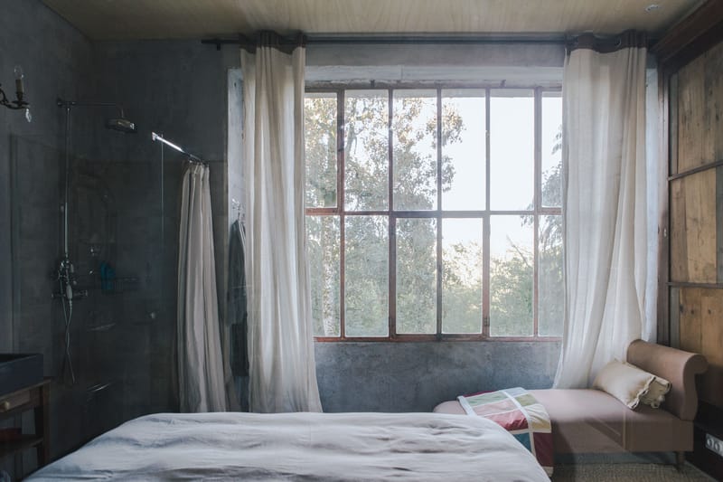 An interior wide shot of a guest bedroom at what appears to be a wedding venue, showing a getting-ready or accommodation space. The room features an industrial-rustic aesthetic with raw concrete walls, a plywood ceiling, and reclaimed wood paneling on the right wall. A double bed with white linen bedding sits in the foreground, and an open-plan glass-enclosed shower with dark tiled walls is visible to the left. Sheer white linen curtains frame a large multi-pane steel-framed window that floods the room with natural light. A blush pink upholstered bench or chaise sits beneath the window with what appears to be a brochure or lookbook placed on it. Potential venue feature image.