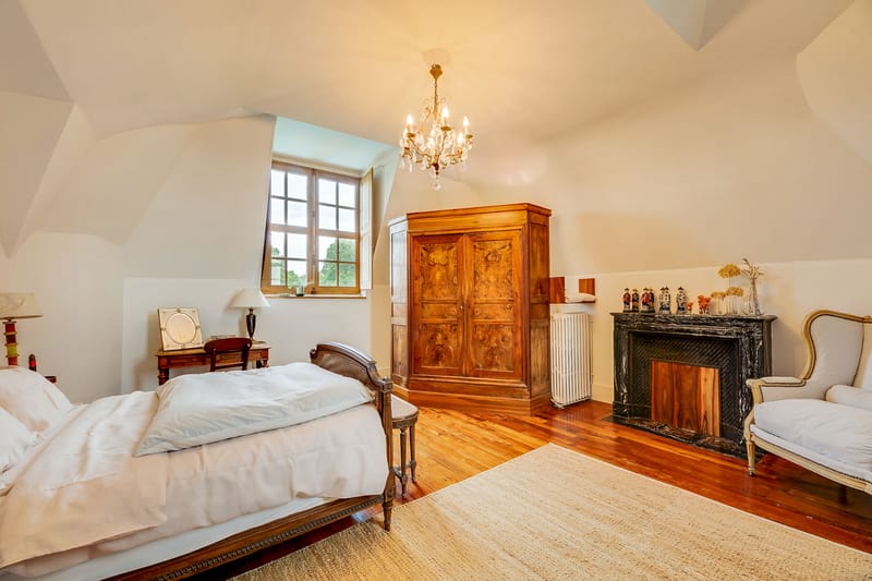 Interior wide shot of a château bedroom, likely used as a bridal suite or guest accommodation. The room features vaulted white ceilings, warm-toned hardwood flooring, and a natural fiber area rug. Furnishings include an antique carved wood bed dressed in white linens, a large burled walnut armoire, a dark mahogany bedside table with a table lamp, and a cream upholstered wingback chair in a French Louis XVI style. A black marble fireplace mantel is decorated with decorative ceramic figurines and a vase of dried ivory stems. A small brass and crystal chandelier hangs from the ceiling, and natural light enters through a multi-pane casement window. No people are present. Potential venue feature image.