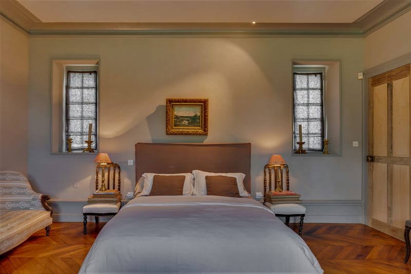 Interior shot of a château bedroom, likely used as a bridal suite or guest accommodation. The room features a large double bed with a taupe upholstered headboard, pale grey-blue bedding, and a mix of ivory and dark brown decorative cushions. Two matching gilt-gold bedside chairs serve as nightstands, each topped with a stack of books and a small lamp with a warm amber shade. Symmetrical tall narrow windows on either side of the bed are dressed with dark sheer linen Roman blinds, and gold candlestick holders sit on the window ledges. A gilt-framed landscape oil painting hangs centered above the headboard on a muted grey-blue wall, and herringbone parquet wood flooring runs throughout. A period-style chaise longue with a grey and white woven fabric is partially visible at the left edge of the frame. The overall styling is classic French château with antique gold accents and a muted grey-brown palette. Wide frontal shot. Potential venue feature image.