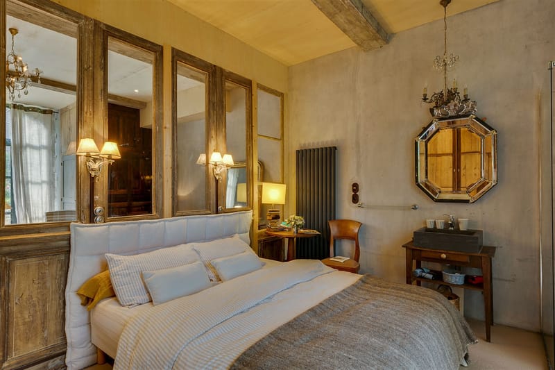 Interior wide shot of a guest bedroom, likely within a French château or manor house venue. The room features a double bed with a white upholstered headboard, grey woven throw, striped pillows, and a mustard yellow accent cushion. The headboard wall is lined with a panel of aged wood-framed mirrors, and two ornate glass wall sconces provide warm ambient lighting. Exposed wooden ceiling beams and bare plaster-finish grey walls contribute to a rustic-meets-refined interior style. A dark slate stone sink sits on a wooden washstand in the corner beneath an octagonal Venetian-style mirror with a decorative candelabra mounted above it. A bedside table holds stacked books, a small floral arrangement, and a table lamp. A tall dark grey vertical radiator is visible against the far wall. Potential venue feature image.