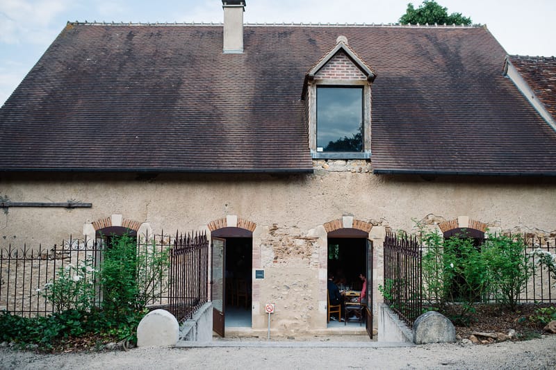 Wide exterior shot of a traditional French rural building featuring a steeply pitched brown tile roof with a central dormer window framed in brick, and a rendered stone facade with arched doorways detailed in red brick and limestone. Two sets of open wooden doors are flanked by wrought iron gates and climbing green shrubs, with rounded stone bollards at the entrance. Through the right-hand doorway, two guests can be seen seated at a wooden table, suggesting a dining or reception space inside. A no-smoking sign is visible near the entrance threshold. Potential venue feature image.