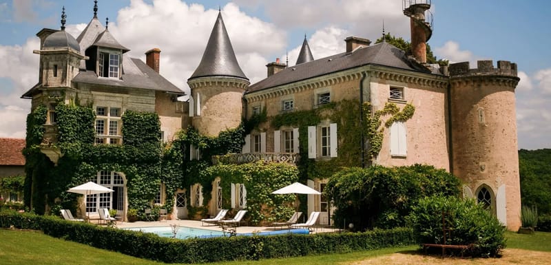 Wide exterior shot of a French château featuring pale stone and pink-toned render walls heavily covered in climbing ivy, with multiple conical-roofed towers, slate grey roofing, white shuttered windows, and crenellated battlements on the right wing. The grounds include a rectangular outdoor swimming pool surrounded by a stone terrace furnished with white sun loungers and two white market umbrellas, with neatly trimmed box hedging bordering the lawn. No people are visible in the image. Potential venue feature image.