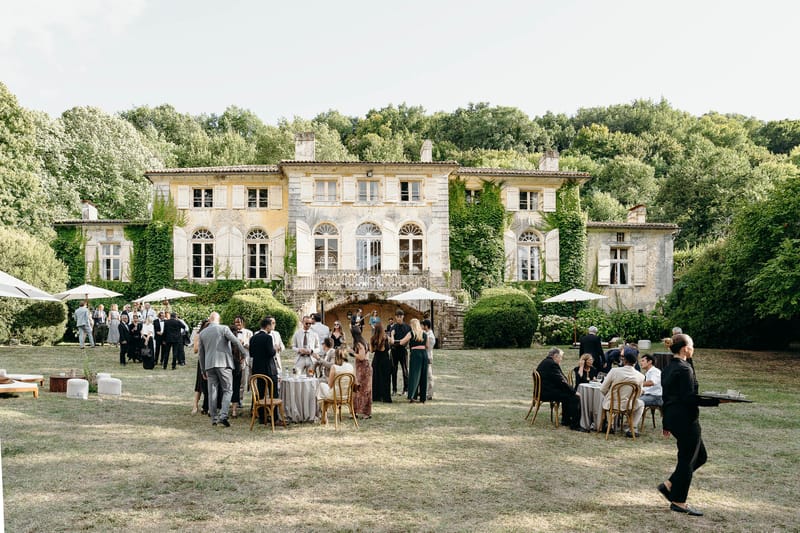 An outdoor cocktail hour is taking place on the lawn in front of a large ivy-covered French manor house with pale limestone facades, arched windows, shutters, and a wrought-iron balcony with exterior stone staircase. Approximately 40–50 guests are mingling across the grounds, seated at round cocktail tables with bentwood chairs or standing in small groups; staff in black uniforms are visible serving guests. White market umbrellas are positioned across the lawn alongside low lounge seating with white cushions on the left side. Guests are dressed in formal attire — suits in grey, navy, and black, alongside floor-length gowns in black, dark green, and floral prints — suggesting a classic, formal wedding aesthetic. The wide-shot composition captures both the full architecture of the venue and the full spread of the cocktail hour activity. Potential venue feature image.