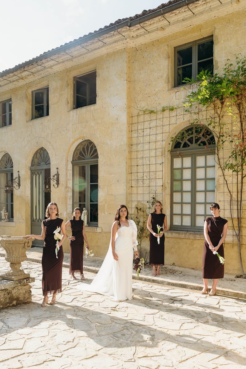 A bridal party portrait taken outdoors on a stone-paved courtyard in front of a two-story French limestone manor with arched windows, grey-green shutters, and a tiled roof. The bride stands at center in a fitted ivory lace gown with a long white cape or train, holding a small bouquet. Four bridesmaids are positioned around her, each wearing a deep burgundy midi-to-maxi sleeveless dress with a high or mock neckline, gold sandals, and carrying single white calla lilies. The styling is modern and minimal with a deliberately muted, dark-and-light contrast palette. A carved stone urn planter is visible to the left. Wide portrait shot in bright natural daylight. Potential venue feature image.