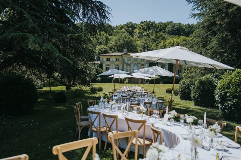 An outdoor wedding reception setup on the grounds of a French manor house, photographed from a slightly elevated wide-angle perspective. Multiple dining tables — a mix of long rectangular and round — are dressed in white linen and set with wine glasses, white taper candles, and small bud vases of white flowers, arranged across a lawn with natural wood cross-back chairs. Large white market umbrellas on copper-toned poles are positioned throughout the setup to provide shade. The decor palette is white and natural wood with a classic, understated style. In the background, a two-storey stone manor house with arched windows and a tiled roof is visible, surrounded by mature trees and manicured hedging. No guests are present — the space is fully set and ready for service. Potential venue feature image.