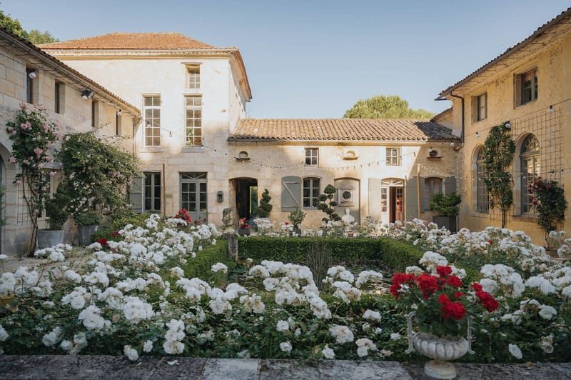 Wide shot of a Provençal-style stone courtyard venue featuring honey-colored limestone buildings with terracotta-tiled roofs, grey-painted shutters, and arched windows. The foreground is filled with densely planted white and red roses in formal garden beds bordered by low boxwood hedging, with a stone urn planter and classical bust sculptures visible among the plantings. Strings of bistro lights are strung across the courtyard between the buildings, and climbing roses in pink and red grow against the façades. No people are present in the image. Potential venue feature image.