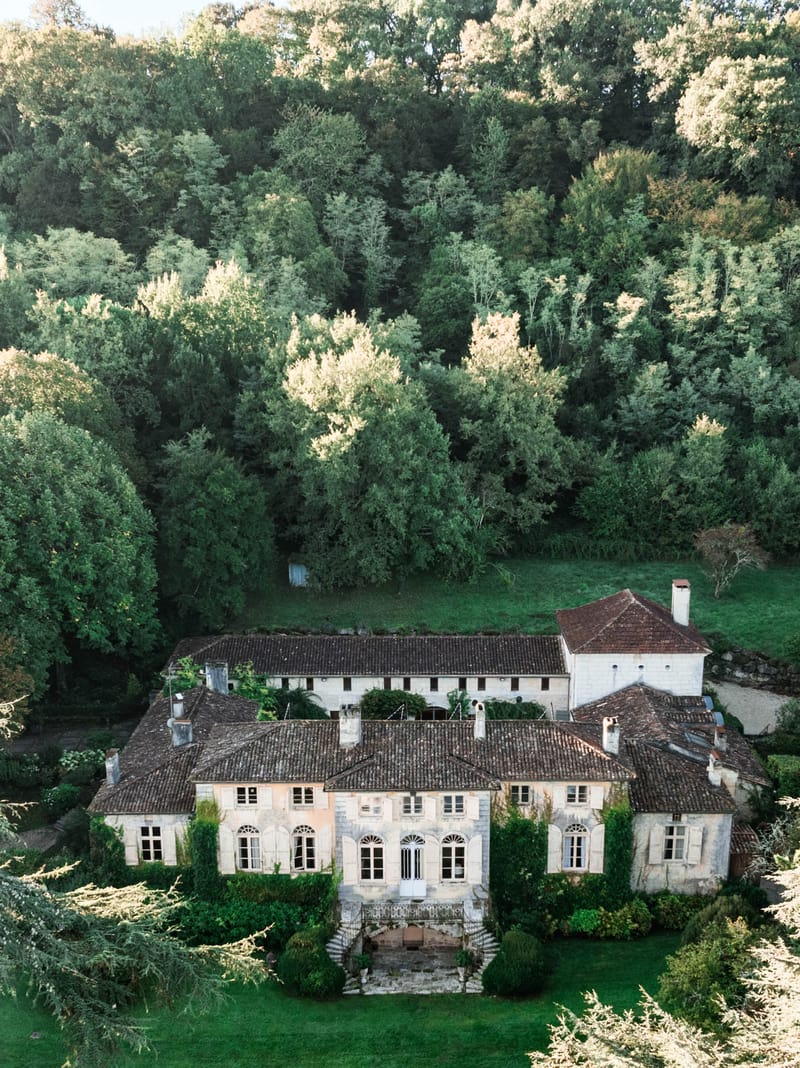 An aerial drone shot of a French country estate, showing a large multi-wing stone manor house with terracotta-tiled roofs, arched fanlight windows, wooden shutters, and a grand front staircase with wrought iron railings. The main building is flanked by outbuildings and a secondary structure with a red-tiled hip roof and chimney, all in pale limestone. Climbing ivy and clipped topiary cover portions of the facade, and a manicured lawn extends in front of the property. The entire estate is set against a dense backdrop of mature woodland. No people are visible in the image. Potential venue feature image.