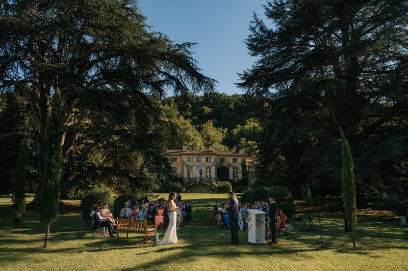 An outdoor wedding ceremony taking place on a wide lawn in front of a large French manor house or château with a terracotta roof, arched windows, and a balcony with exterior staircases. The bride, wearing a white fitted gown with ruffle or lace detailing, stands facing the groom, who is dressed in a dark suit, with an officiant at a white pedestal lectern to the right. Approximately 50–70 seated guests are arranged in rows on the lawn, with a wooden bench visible at the front left. The wide-angle shot frames the ceremony between large cedar trees and tall cypress trees, with the manor facade visible in the background mid-distance. The ceremony has a classic, unfussy outdoor style with no visible floral arches or decor beyond the white lectern. Potential venue feature image.