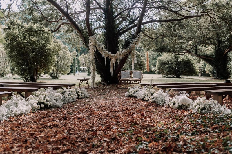An outdoor ceremony setup photographed before the ceremony, with no people present. The altar is centered beneath a large tree, with a floral garland of white roses and baby's breath draped in a crescent arc across the trunk, accented with hanging macramé or fringe details in a boho style. A rattan loveseat with white cushions serves as the couple's seat, flanked by a small wooden side table. Dark wood benches are arranged in two rows forming an aisle, which is lined on both sides with loose clusters of white hydrangeas, white roses, and baby's breath arranged directly on the ground. The aisle floor is covered in rust-colored fallen leaves. The overall decor palette is all-white florals against natural wood tones, consistent with a rustic-boho aesthetic. Wide establishing shot taken from the back of the aisle looking toward the altar. Potential venue feature image.