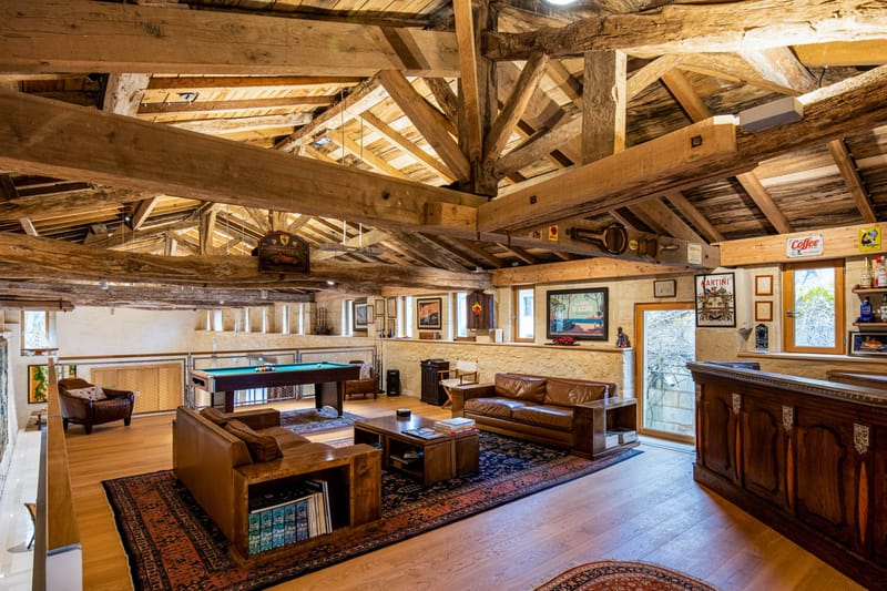 Interior wide shot of a rustic venue games room or lounge space, featuring exposed heavy timber roof beams with a high vaulted ceiling and light oak plank flooring. The room is furnished with brown leather sofas arranged around a dark wood coffee table on a deep red and navy Persian-style rug, with a green-baize pool table positioned behind the seating area. To the right is a dark wood bar counter with vintage enamel signs including Martini and Coffee branding mounted on the stone walls, alongside framed vintage travel posters including a Côte d'Azur print. No people are present in the image. The overall decor palette combines warm amber wood tones, dark brown leather, and deep red textiles in a rustic-meets-vintage style. Potential venue feature image.