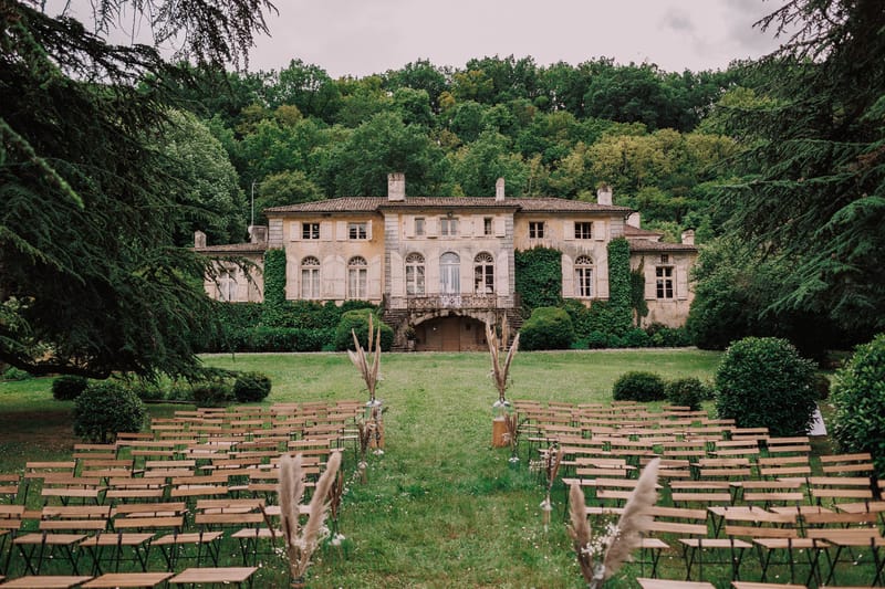 An outdoor wedding ceremony setup on the lawn of a large French manor house or château, with no guests or couple present — the space is arranged and awaiting the ceremony. Rows of light wood folding chairs with black metal frames are arranged in two sections separated by a central grass aisle. The aisle is lined with dried pampas grass and small glass bud vases, along with taller arrangements of dried grasses and delicate white wildflowers placed on wooden pedestals at intervals. The décor palette is natural and earthy, leaning toward a boho-rustic style with dried botanical elements rather than fresh floral arrangements. The manor building in the background is a two-storey stone structure with ivy-covered facades, arched windows with shutters, a wrought-iron balcony, and a tiled roof. Wide shot taken from behind the guest seating looking toward the house. Potential venue feature image.