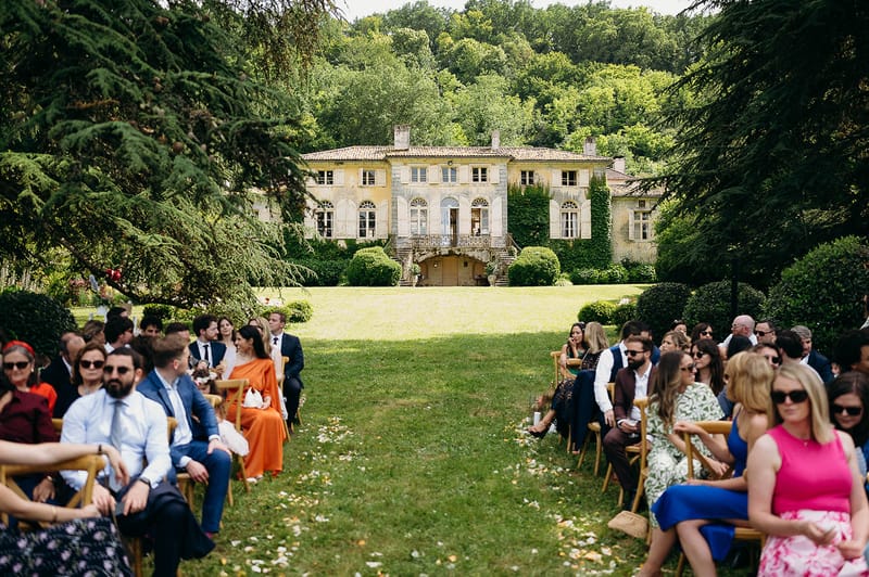An outdoor wedding ceremony taking place on the grounds of a large French château, with guests seated on wooden folding chairs arranged in two rows on either side of a central grass aisle scattered with flower petals. Approximately 40–50 guests are visible, dressed in colorful summer attire including an orange floor-length dress, a blue dress, a pink dress, and various suits and blazers. The ceremony appears to be in progress with guests facing away from the camera toward the front of the aisle. The château in the background is a two-storey stone manor house with a terracotta tiled roof, arched windows, ivy-covered walls, a wrought-iron balcony, and manicured box hedging at its base. The wide-angle shot is taken from behind the seated guests, framing the full façade of the property at the end of the aisle. Potential venue feature image.
