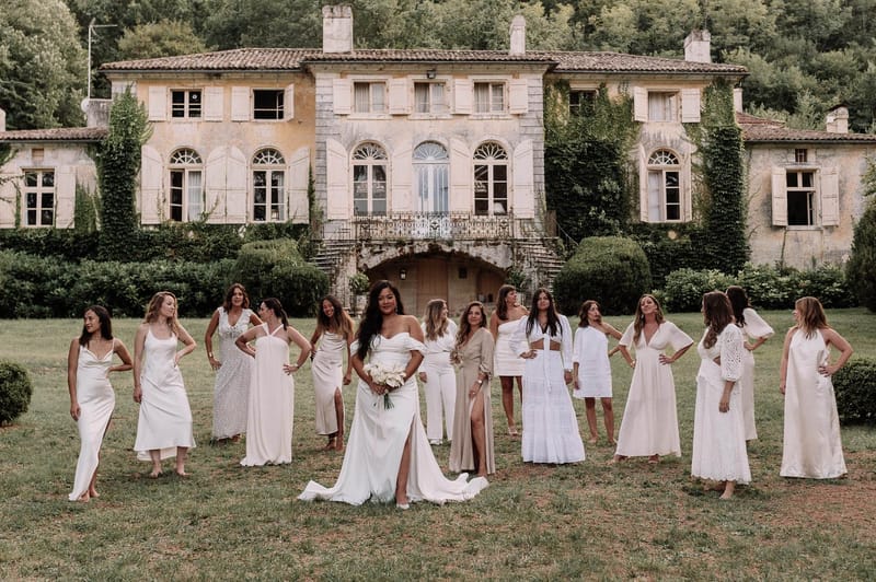 A bridal party group portrait taken outdoors on the lawn of a large French manor or bastide, featuring the bride and approximately twelve bridesmaids arranged in a loose arc. The bride stands at center, wearing an off-the-shoulder white gown with a front slit and train, holding a bouquet of white calla lilies or tulips. The bridesmaids wear a mix-and-match palette of white, ivory, and champagne-beige outfits in varied styles including satin slip dresses, lace midi dresses, a two-piece crop top set, a sheer coverup, and a structured mini dress, creating a cohesive but individualized look. The venue is a multi-story Provençal-style manor with arched windows, white shutters, ivy-covered facades, a wrought-iron balcony, and a grand stone entrance staircase. The overall styling theme is modern boho with a white and neutral color palette. Wide shot composition.