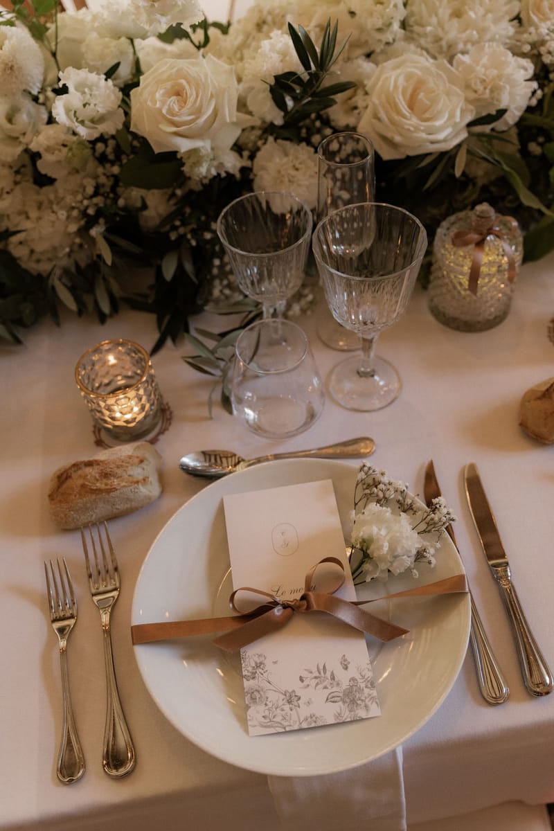 A close-up detail shot of a wedding reception place setting on a white linen tablecloth. The white plate holds a menu card labeled 'Le menu' tied with a warm brown satin ribbon bow, accompanied by a small sprig of white gypsophila and white flowers as a decorative accent. The menu card features a floral botanical print in sepia tones. Silver cutlery is arranged on either side of the plate — two forks to the left and a knife and additional utensil to the right — with a spoon placed above the plate. Three faceted crystal glasses are positioned above the setting, and two small mercury glass votive candle holders with lit tea lights flank the place setting. A bread roll sits to the upper left of the plate. The centerpiece visible in the background is a lush, low arrangement of ivory roses, white carnations, white hydrangea, gypsophila, and olive branch foliage. The overall decor palette is ivory, white, and warm gold, with a classic and refined styling approach.