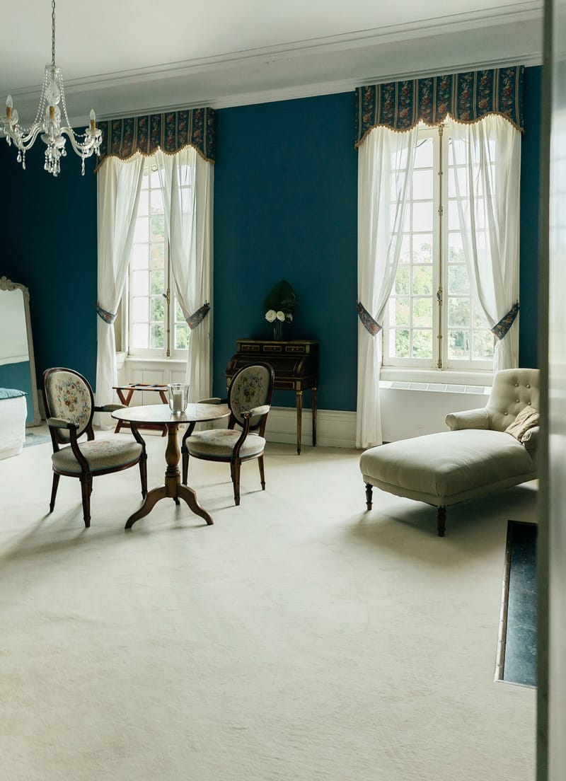 An interior room shot of what appears to be a château or manor house, showing a classical French-style sitting room with no people present. The walls are painted deep teal blue and are decorated with two tall white-framed windows dressed with white sheer curtains and floral-patterned navy and gold valances. The room features a crystal chandelier, a round dark wood pedestal table surrounded by three antique balloon-back chairs upholstered in floral needlepoint fabric, a cream tufted chaise longue, a dark wood roll-top writing desk with a small white floral arrangement, and a large ornate gilt-framed mirror partially visible on the left. The cream carpet and white crown molding contrast with the deep teal walls. Wide shot taken from a doorway. Potential venue feature image.