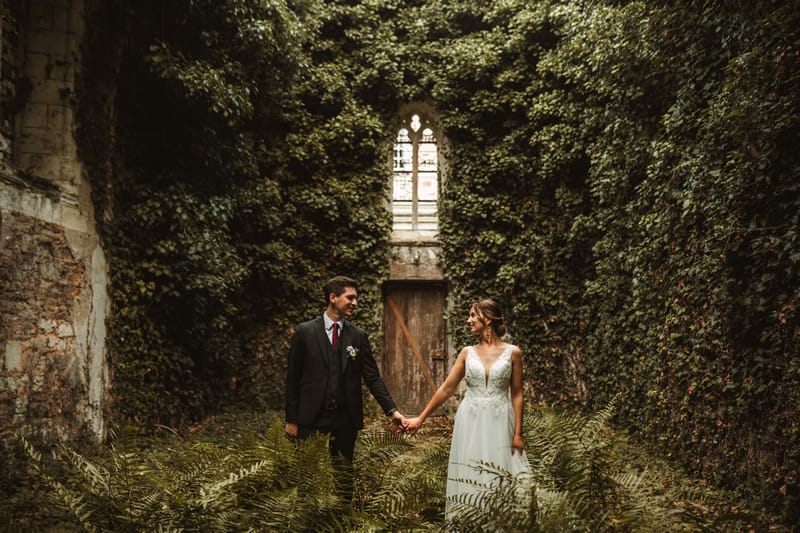 A couple portrait taken outdoors in what appears to be a ruined or overgrown courtyard, with ivy-covered walls completely surrounding them and a weathered wooden door topped by a Gothic arched window in the background. The bride wears a white sleeveless gown with a deep V-neckline and lace bodice detailing in a bohemian style, with her hair pinned up and drop earrings; the groom wears a dark charcoal suit with a burgundy tie and a white floral boutonnière. The two are holding hands and looking at each other while standing among ferns growing at ground level. The image is shot in a mid-wide portrait format with moody, dark-toned natural lighting that emphasizes the overgrown, atmospheric setting.