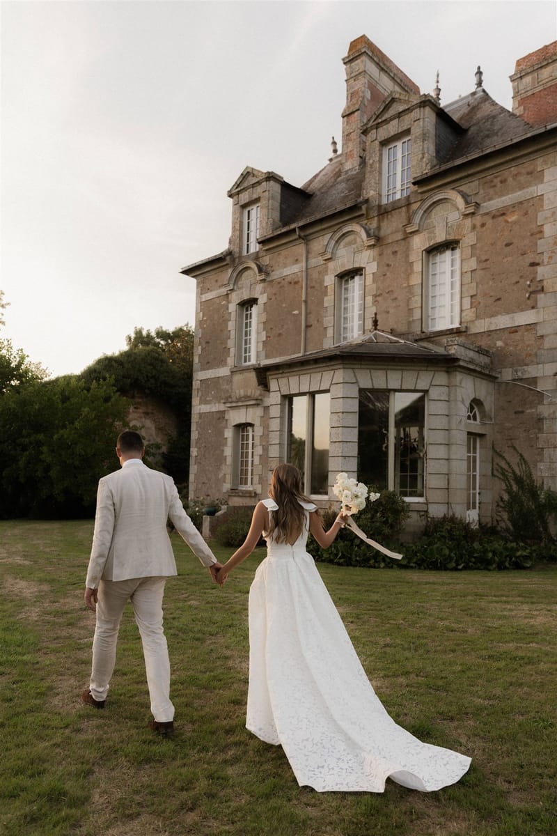 A couple portrait shot from behind showing the bride and groom walking hand-in-hand across a lawn toward a multi-story French château in warm evening light. The groom wears a light beige linen suit with brown shoes, and the bride wears a white gown with a low open back, bow shoulder straps, a textured or lace-patterned full skirt, and a long trailing train. She carries a loose bouquet of white flowers, including what appear to be white roses or peonies, with trailing ivory ribbon. The wide shot frames the full façade of the stone château, which features arched windows, ornate stone detailing, a bay window extension, and a mansard-style roofline. Potential venue feature image.
