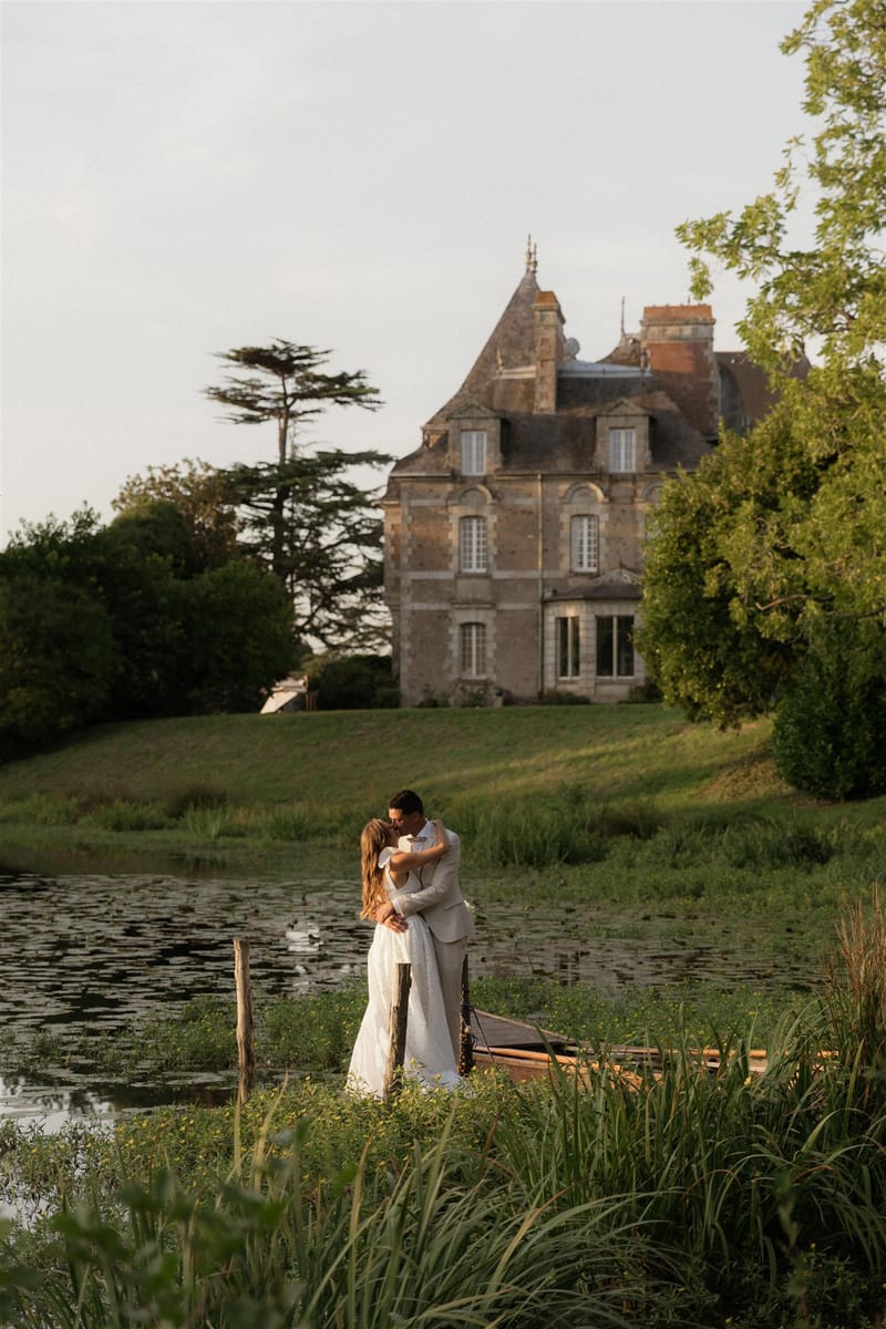 A couple portrait taken outdoors at golden hour, with the bride and groom embracing and kissing on the bank of a lily pad-covered pond. The bride wears a white fitted gown and the groom is dressed in a light beige suit. A small wooden rowboat is moored at the water's edge behind them. The setting is the grounds of a French château — a multi-story stone manor house with steeply pitched slate roofs, arched windows, and classical architectural detailing — visible on a hill directly behind the couple. The composition is a wide shot taken from across the pond, framing the couple in the middle ground with the château as a backdrop. Warm late-afternoon light bathes the scene. Potential venue feature image.