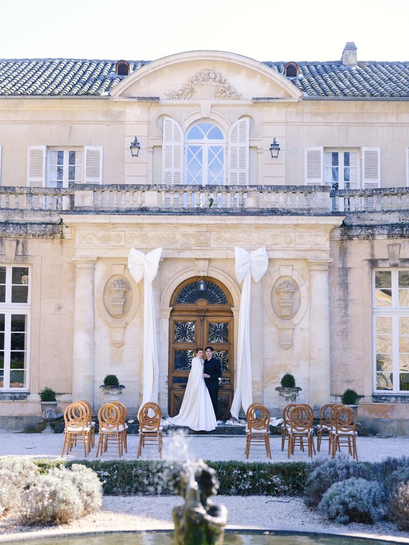 Couple stands at château door under oversized ivory bows with cane ceremony chairs on gravel courtyard