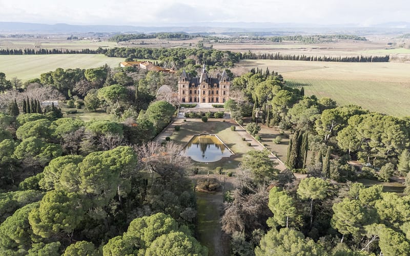 Aerial view of a terracotta-roofed chateau surrounded by manicured gardens, cypress trees, and rolling countryside.