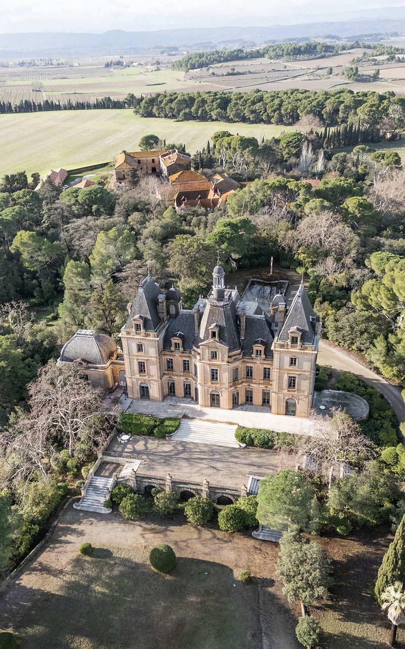 Aerial view of 19th-century château with mansard roof surrounded by cypress trees and manicured grounds.
