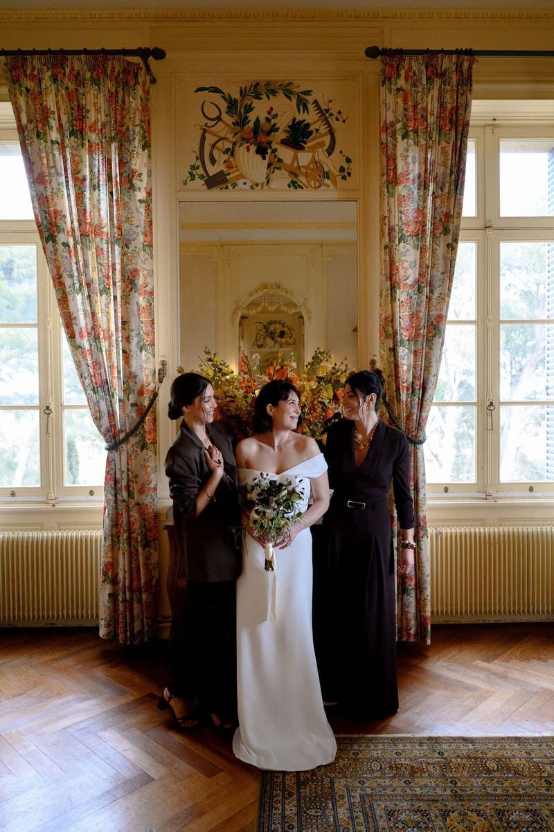 Bride and two friends before tall windows and painted overdoor panel in Château du Terral's Claude suite
