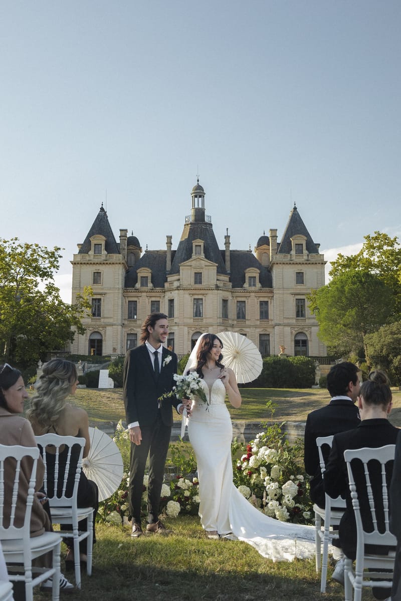 Outdoor wedding ceremony on the lawn before Château du Terral, guests holding parasols