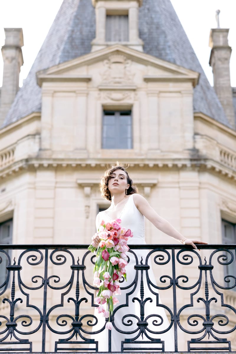 Bride with a cascading pink bouquet on an iron balcony below a Château du Terral turret