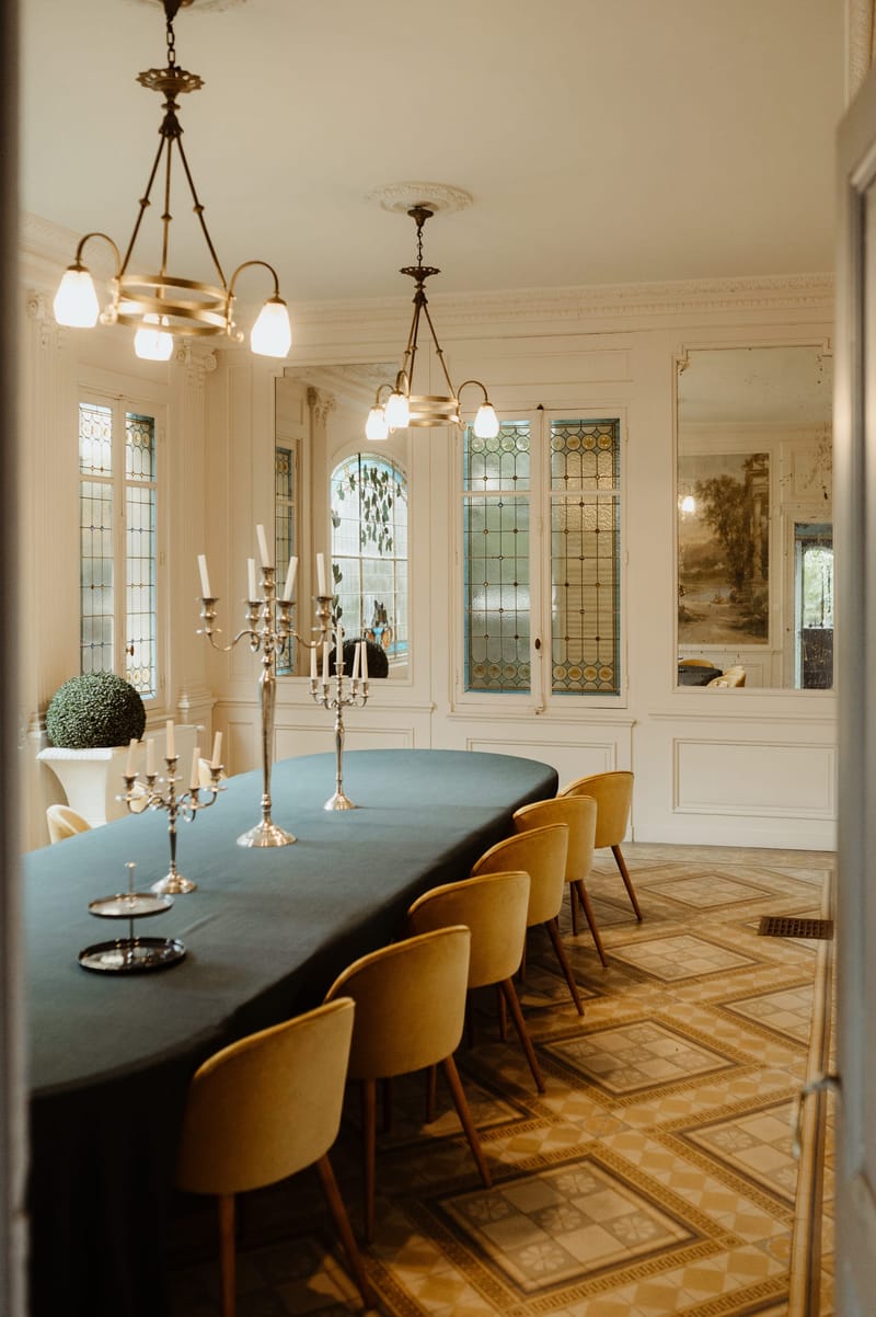 Formal dining room with stained-glass windows, chandeliers and a long table set with mustard velvet chairs