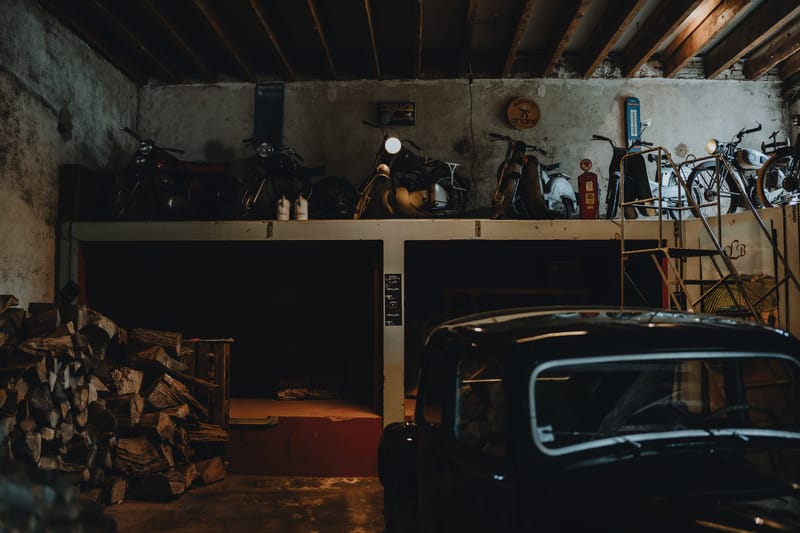 Vintage motorcycles and a classic car displayed on a raised platform in a stone outbuilding