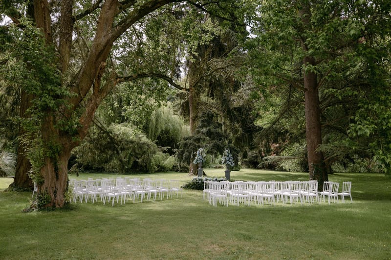 Rows of white ceremony chairs set beneath mature trees on the lawn