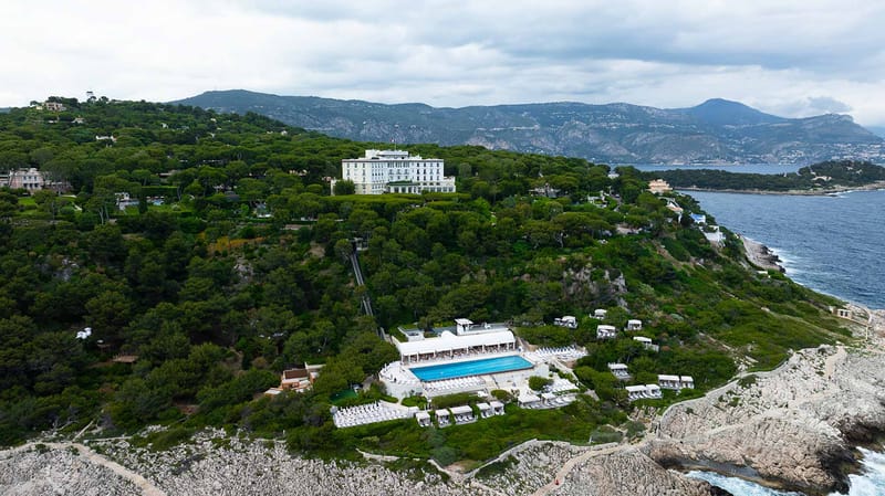 Aerial wide shot of a large white Belle Époque-style villa and its grounds situated on a wooded coastal headland on the French Riviera. The property features a grand multi-storey white building with a classical facade set among dense pine trees, connected via a funicular track to a lower pool and beach club terrace. The lower level includes a large rectangular outdoor swimming pool with white sun loungers arranged around it, white cabanas, and a white-roofed pavilion structure. The architecture and decor palette throughout is entirely white. The property sits directly above rocky coastal cliffs with the Mediterranean Sea visible to the right and mountain ranges in the background. No people or wedding-specific elements are visible at this scale. Potential venue feature image.