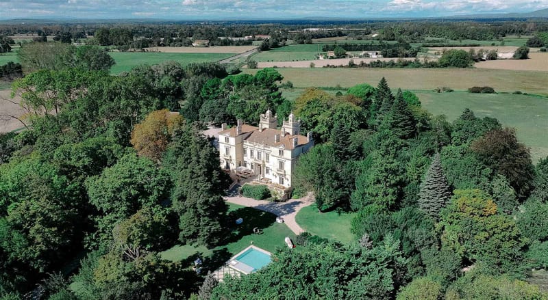 Aerial drone shot of a French château set within extensive private grounds, showing the full property from above. The building is a cream-rendered, multi-storey manor house with a terracotta tile roof, ornate roofline details including dome-topped turrets, and symmetrical facade with tall arched windows. A paved forecourt with a curved driveway sits at the front of the building, and a terrace with outdoor furniture is visible along the main facade. A rectangular swimming pool with blue water is visible in the lower portion of the grounds, surrounded by a manicured lawn. The property is entirely surrounded by a dense parkland of mature trees, with open agricultural fields and countryside extending to the horizon beyond the estate boundary. Potential venue feature image.